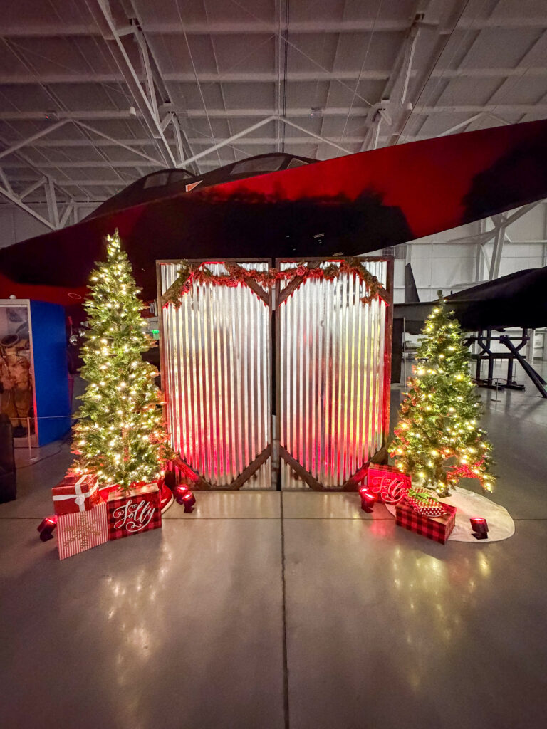 Holiday-themed photo backdrop with corrugated metal panels, decorated Christmas trees, wrapped gifts, and festive lighting inside the Hill Aerospace Museum.