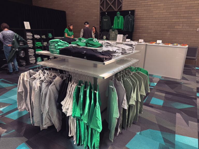 Conference merchandise display featuring branded hoodies, T-shirts, hats, and apparel neatly arranged on racks and shelving for attendees to browse.