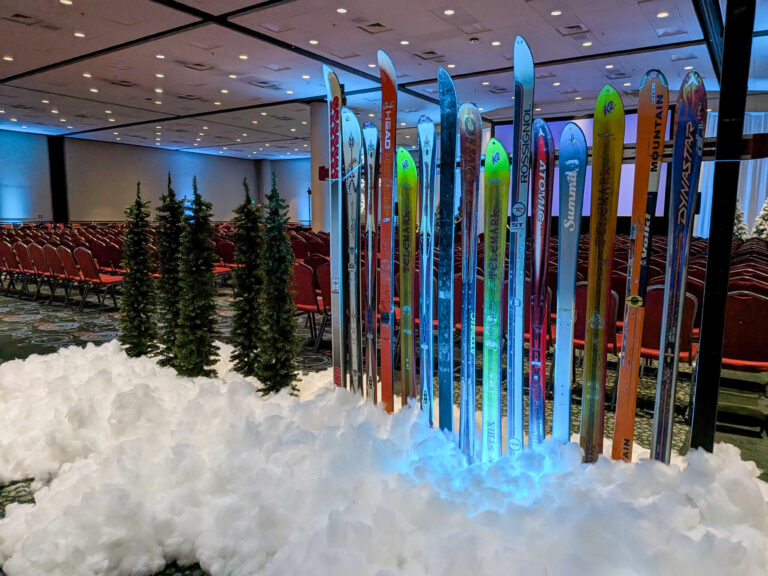 Conference ballroom featuring vintage skis, artificial snow, evergreen trees, and winter décor leading toward a presentation stage.