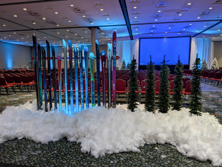 Conference ballroom featuring vintage skis, artificial snow, evergreen trees, and winter décor leading toward a presentation stage.