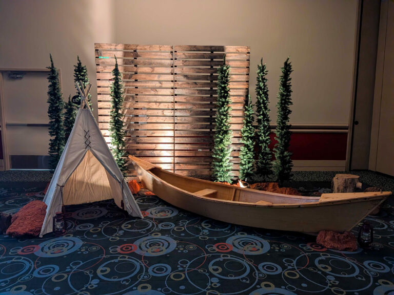Rustic camp-themed event display featuring a wooden canoe, teepee, evergreen trees, pallet backdrop, and lanterns for a conference photo opportunity.