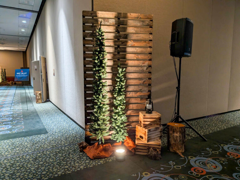 Rustic event vignette with wooden pallet backdrop, illuminated evergreen trees, lantern, crates, and woodland décor for a conference or holiday event.