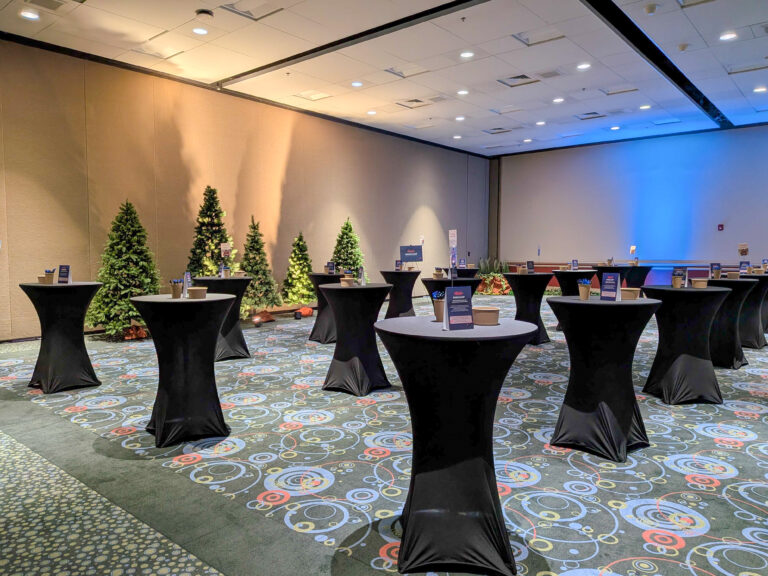 Corporate cocktail reception featuring black spandex cocktail tables, evergreen trees, and warm accent lighting for a winter-themed event.
