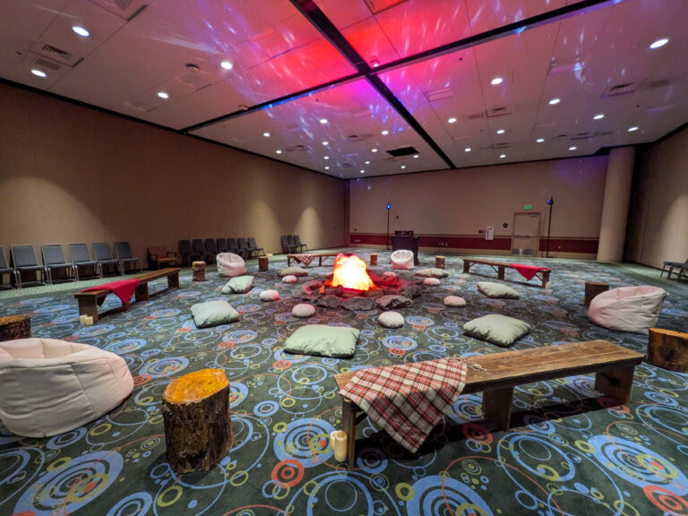 Indoor campfire lounge featuring an LED fire pit, rustic benches, tree stump seating, bean bags, and cozy décor for a corporate event.