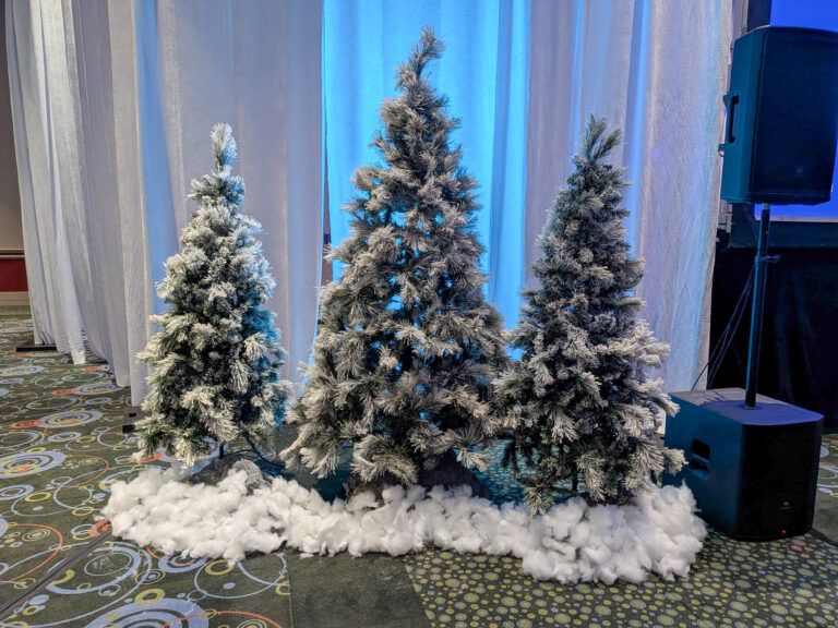 Flocked evergreen trees with faux snow displayed against white drape and blue uplighting for a winter-themed corporate event.