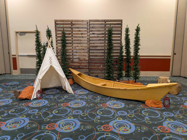Rustic event photo backdrop featuring a wooden canoe, teepee tent, evergreen trees, pallet wall, and woodland décor for a themed corporate event.