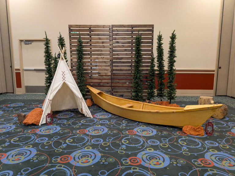 Rustic event photo backdrop featuring a wooden canoe, teepee tent, evergreen trees, pallet wall, and woodland décor for a themed corporate event.