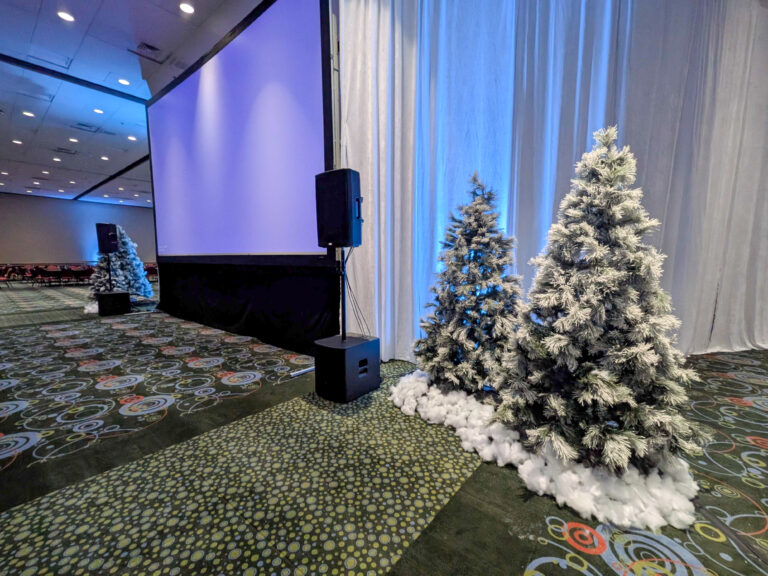 Flocked Christmas trees with faux snow, white drape backdrop, and blue uplighting framing a projection screen at a winter corporate event.