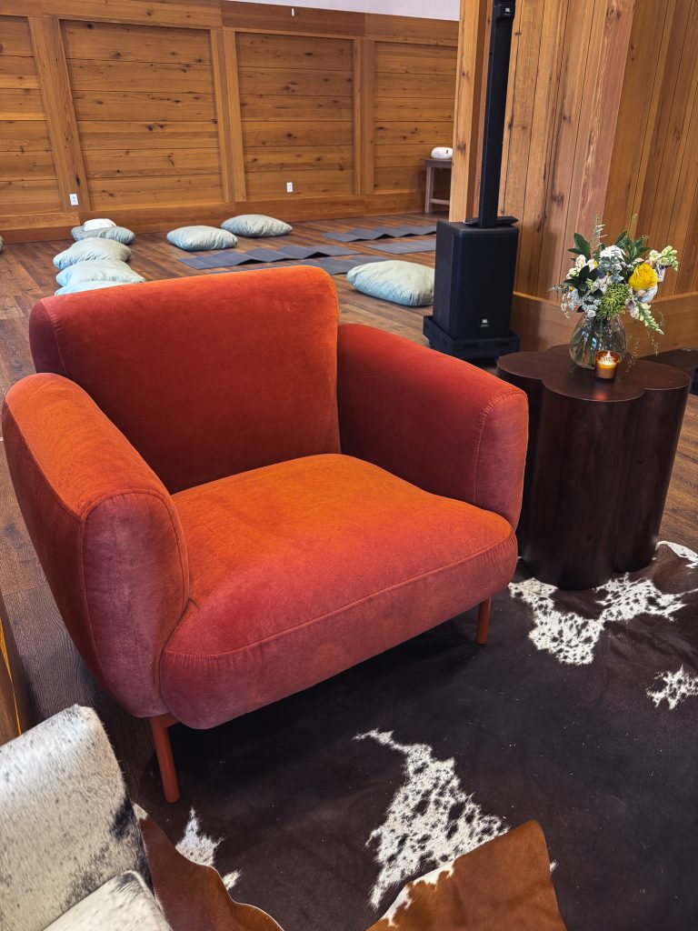 Rust-colored velvet lounge chair paired with a dark wood clover side table, floral arrangement, and cowhide rug.
