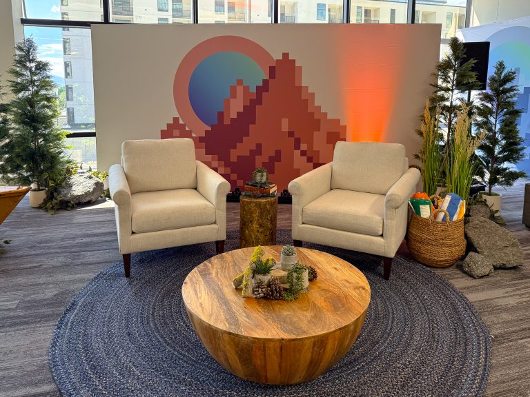 Camp-inspired lounge with cream armchairs, a round wood coffee table, evergreen trees, rocks, and a custom mountain backdrop.