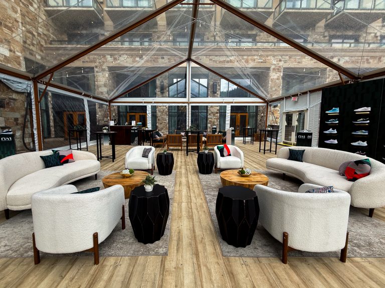 Modern event lounge furniture featuring curved white sofas, upholstered chairs, wood coffee tables, and black accent tables beneath a clear tent at St. Regis Deer Valley.