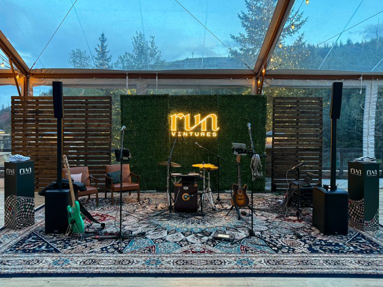 Illuminated Run Ventures logo on a green boxwood backdrop behind a live music stage with guitars, drums, and speakers at St. Regis Deer Valley.