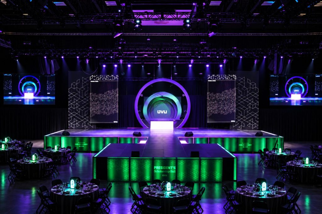 Wide view of the portal stage with a circular LED display, floating side screens, outboard video screens, and a backlit green skirt above the centerpiece tables at the UVU President's Scholarship Ball 2026 in Orem, Utah